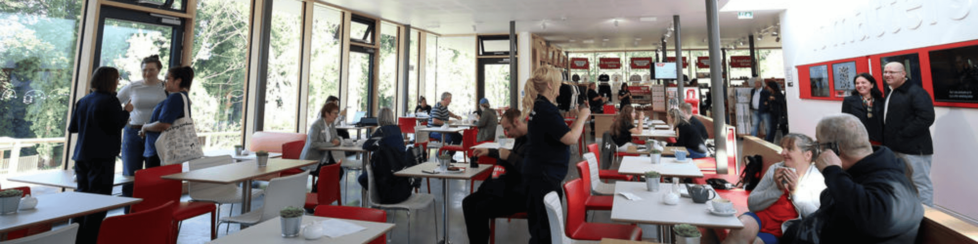 An indoor cafe with huge windows in the sunlight with white tables and red chairs. People are sat eating on this tables and smiling.