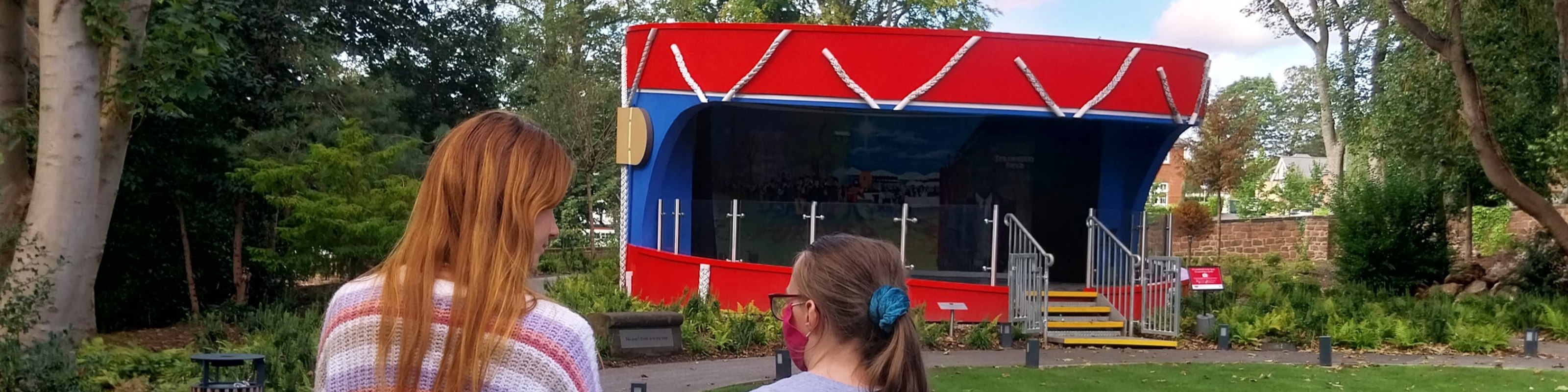 Two people stand facing a red and blue bandstand in gardens at Strawberry Field